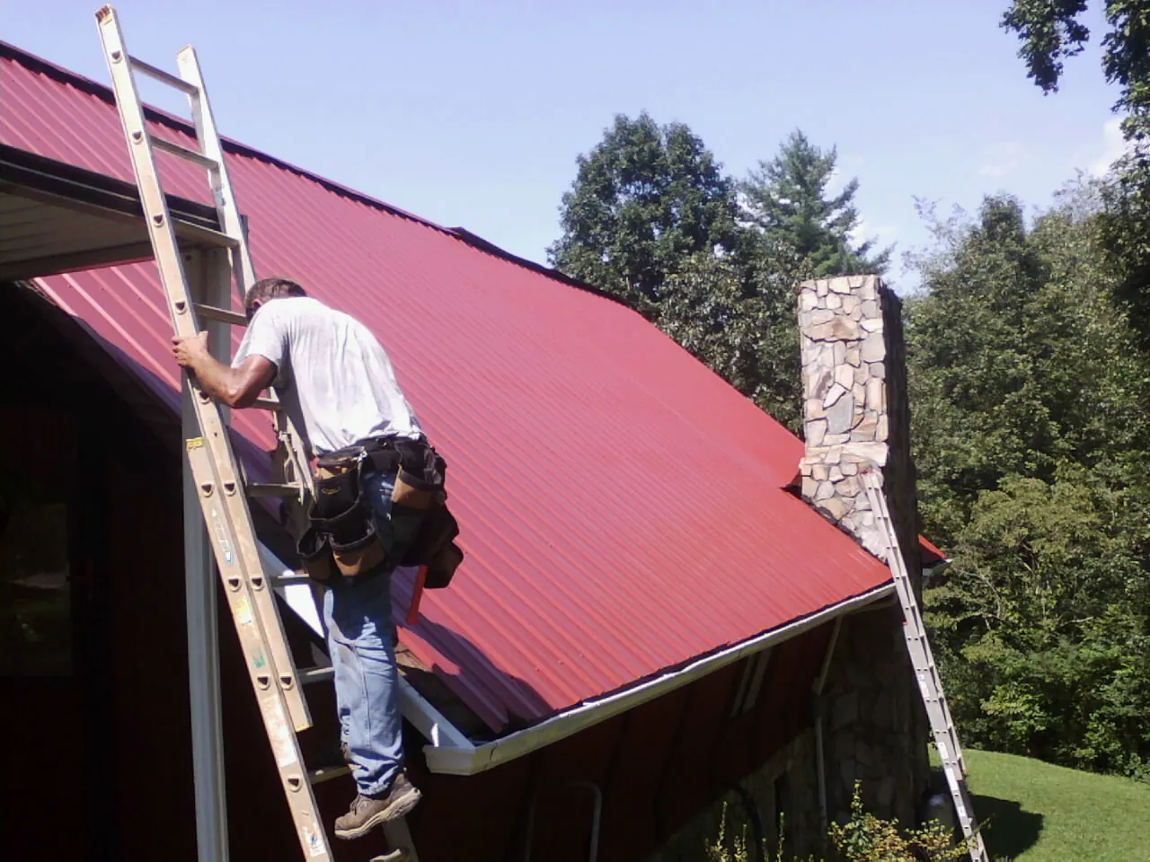 Roofer on a ladder beside a red metal roof and stone chimney — Soffit & Fascia Repair by Roofing Brothers in Hooversville